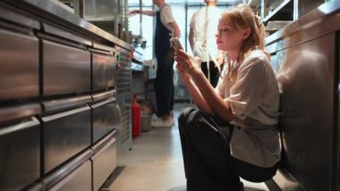 Dost canlısı sarışın kadın şef restoranın mutfağında telefonla mesaj atıyor.