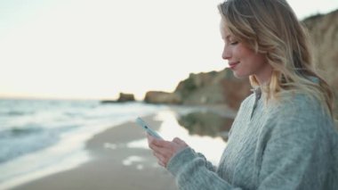 Neşeli sarışın kadın deniz kenarındaki kumsalda cep telefonunda yazı yazıyor.