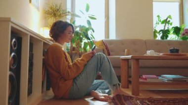 Kahkahalar atan, gözlüklü esmer kadın cep telefonuyla evde yerde yazı yazıyor.