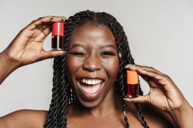 Gri arka planda izole edilmiş ojelerle poz verirken üstsüz siyah kadın göz kırpıyor.