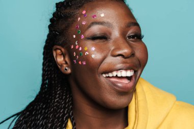 Yüzünde çıkartmalar olan genç siyahi bir kadın mavi arka planda izole edilmiş kameraya göz kırpıyor.