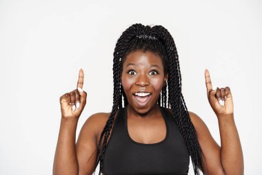 Siyahi genç kadın şaşkınlığını dile getiriyor ve beyaz arka plan üzerinde parmaklarını yukarı kaldırıyor.