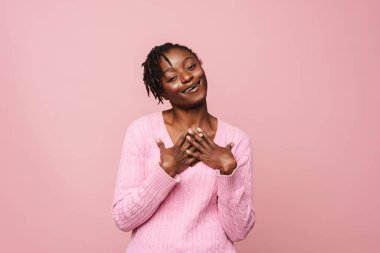 Gülümseyen ve göğsünde el ele tutuşan genç bir kadın pembe arka planda izole edilmiş.