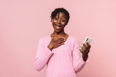 Pembe arka planda izole edilmiş cep telefonu kullanan siyah, şaşırmış bir kadın.