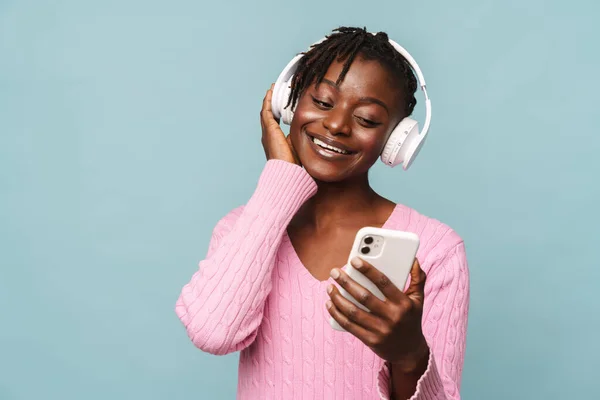 アフリカ系アメリカ人の若い女性は 青い背景に隔離された携帯電話やヘッドフォンを使用して笑顔 — ストック写真