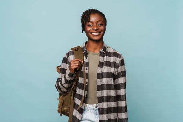 穿着格子呢衬衫和背包的非洲裔美国女青年对着蓝色背景的相机笑着 — 图库照片