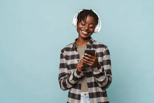 青い背景に隔離されたヘッドフォンや携帯電話を使用して再生シャツでアフリカ系アメリカ人の若い女性 — ストック写真