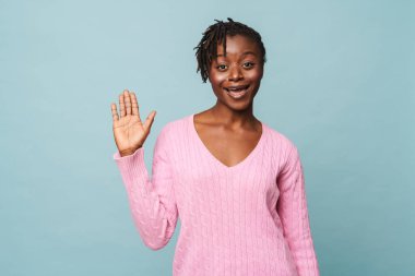 Afro-Amerikan mutlu kadın gülümsüyor ve elini mavi arka planda izole bir şekilde karşılama hareketi yapıyor.