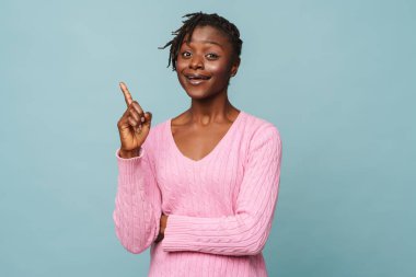 Afro-Amerikalı mutlu kadın gülümsüyor ve mavi arkaplan üzerinde izole edilmiş boşluğu işaret ediyor.