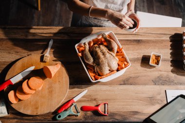 Evdeki mutfakta tavuk pişirirken önlük takan genç bir kadın tablet bilgisayar kullanıyor.