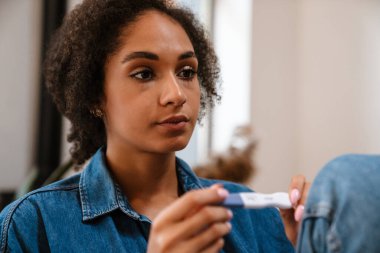 Genç siyahi kadın hamilelik testini evdeki kanepede oturarak yaparken üzülüyor.