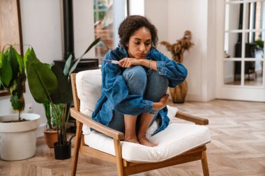 Genç siyahi kadın hamilelik testini evdeki kanepede oturarak yaparken üzülüyor.