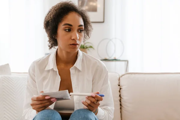 年轻的黑人妇女坐在家里的沙发上进行妊娠测试时很不高兴 — 图库照片