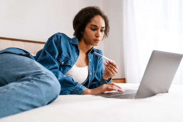Young Black Woman Holding Pregnancy Test Using Laptop While Lying — Stock Photo, Image