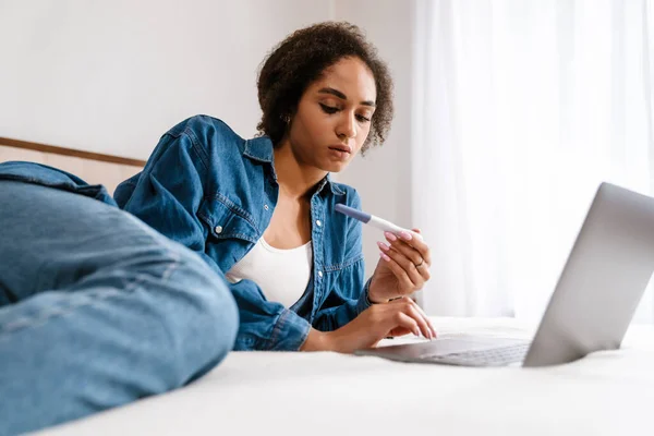 Young Black Woman Holding Pregnancy Test Using Laptop While Lying — Stock Photo, Image
