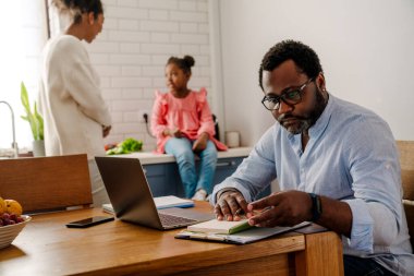 Kadın ve küçük kız mutfakta kahvaltı hazırlarken, Afrikalı Amerikalı bir adam dizüstü bilgisayarla çalışıyor.