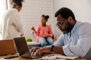 Kadın ve küçük kız mutfakta kahvaltı hazırlarken, Afrikalı Amerikalı bir adam dizüstü bilgisayarla çalışıyor.
