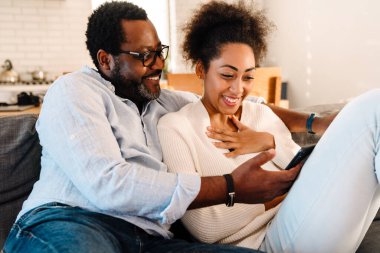 Afro-Amerikalı yetişkin kadın ve erkek evde koltukta otururken gülümsüyor ve cep telefonu kullanıyor.