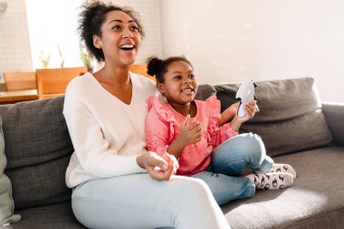 Afrikalı Amerikalı bir kadın ve küçük bir kız kanepede oturuyor ve evde kontrolörle oyun oynuyorlar.