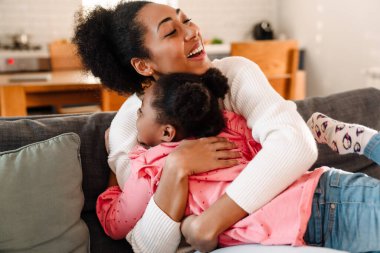 Afro-Amerikan mutlu yetişkin kadın gülümseyerek kızını kucaklıyor.