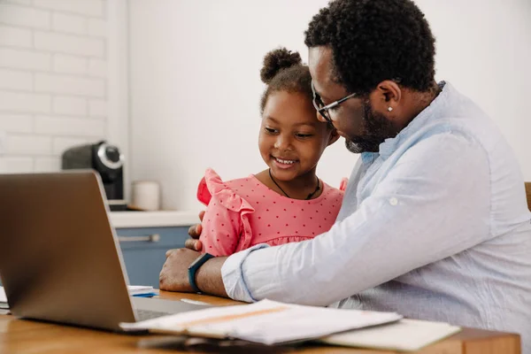 African american adult man hugging his little daughter while working on laptop at home