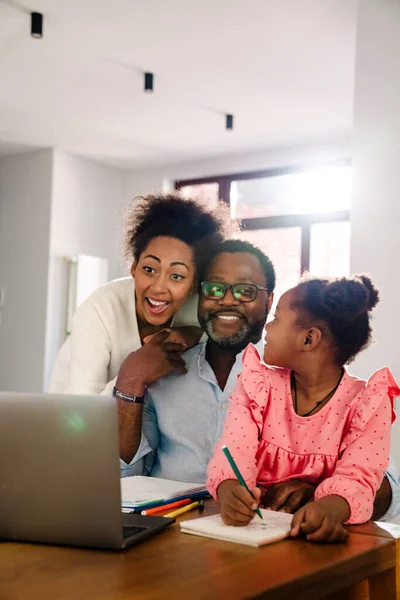 African american family with little girl smiling and using laptop while sitting by table at home