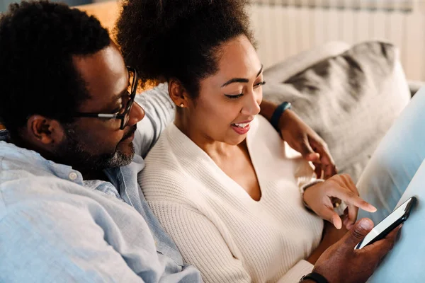 アフリカ系アメリカ人の大人の女性と男の笑顔と携帯電話を使用しながら 自宅でソファに座って — ストック写真