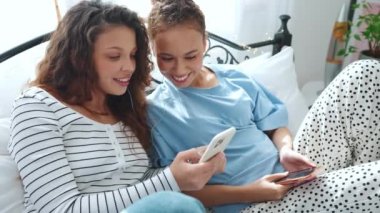 Neşeli kız arkadaşlar cep telefonlarına bakar ve kulaklıkla yatakta müzik dinlerler.