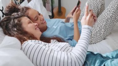 Evdeki yatakta konuşan ve cep telefonuna bakan iki kadın.