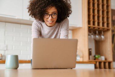 Evde dizüstü bilgisayar kullanan kıvırcık saçlı ve gözlüklü Afro-Amerikan genç kadın.