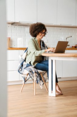 Evde dizüstü bilgisayar kullanan kıvırcık saçlı ve gözlüklü Afro-Amerikan genç kadın.