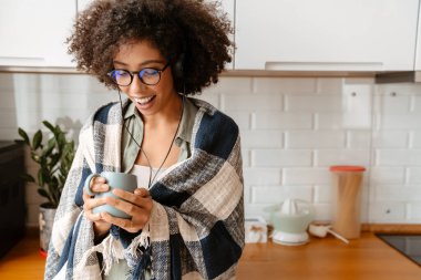 Afro kıvırcık saçlı, evde kahve içerken kulaklık kullanan Afro-Amerikan genç bir kadın.