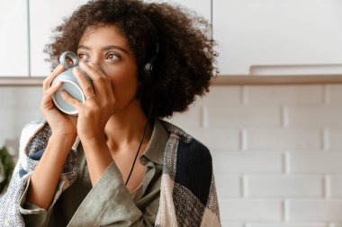 Afro kıvırcık saçlı, evde kahve içerken kulaklık kullanan Afro-Amerikan genç bir kadın.