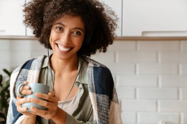 Afro kıvırcık saçlı, evde kahve içerken kulaklık kullanan Afro-Amerikan genç bir kadın.