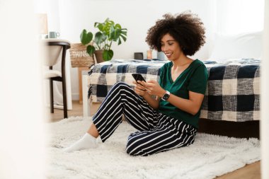 Afro kıvırcık saçlı, gülümseyen ve cep telefonu kullanan Afro-Amerikan genç kadın.