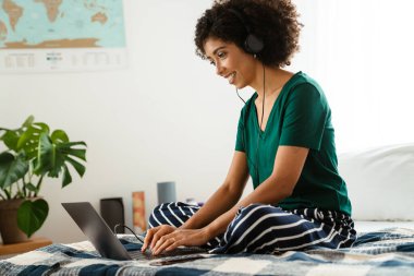 Afro kıvırcık saçlı, yatakta oturan ve evde dizüstü bilgisayar kullanan Afro-Amerikan genç kadın.