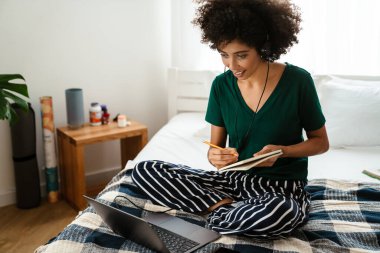 Afro-Amerikalı genç bir kadın yatakta oturup dizüstü bilgisayar kullanırken not alıyor.