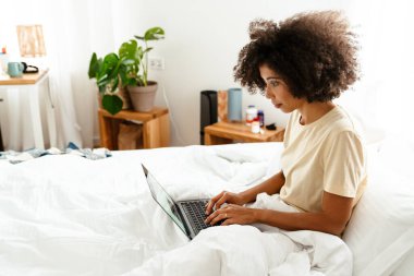 Kıvırcık saçlı, yatakta yatan ve evde dizüstü bilgisayar kullanan Afrikalı Amerikalı genç bir kadın.