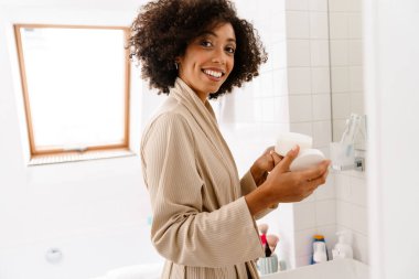 Kıvırcık saçlı, gülümseyen ve yüz kremini banyoda tutan Afro-Amerikan genç kadın.