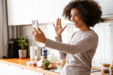 Evde cep telefonu kullanırken kıvırcık saçlı, gülümseyen Afrikalı Amerikalı genç bir kadın.