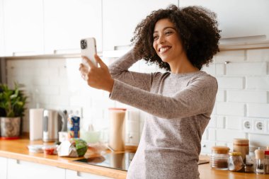 Evde cep telefonu kullanırken kıvırcık saçlı, gülümseyen Afrikalı Amerikalı genç bir kadın.