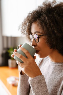Afro kıvırcık saçlı, sabah evde kahve içen Afro-Amerikalı genç bir kadın.