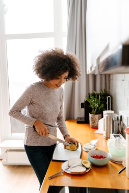 Afro kıvırcık saçlı Afro-Amerikalı genç bir kadın sabah kahvaltısını evde hazırlıyor.