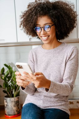Evde cep telefonu kullanırken kıvırcık saçlı, gülümseyen Afrikalı Amerikalı genç bir kadın.
