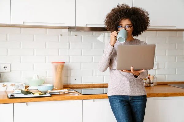 アフリカ系アメリカ人の若いです女性ともに巻き毛コーヒーを飲みながら使用ノートパソコンでホーム — ストック写真