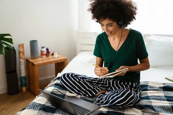 非洲裔美国女青年坐在床上做笔记 在家使用笔记本电脑 — 图库照片