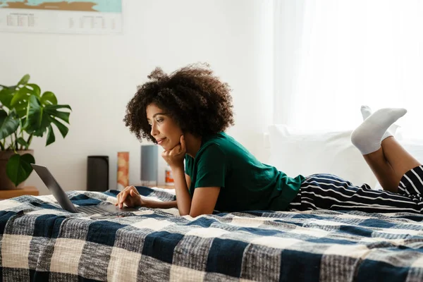 头发卷曲的非洲裔美国女青年躺在床上 在家里用笔记本电脑 — 图库照片