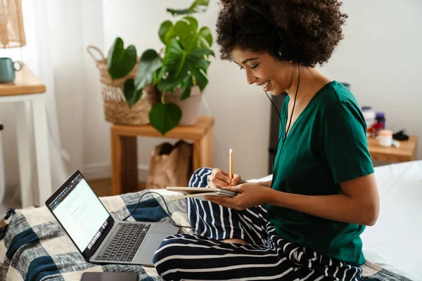 非洲裔美国女青年坐在床上做笔记 在家使用笔记本电脑 — 图库照片