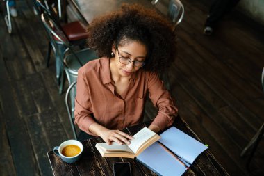 Afro saç stili olan genç siyahi bir kadın. Kapalı kafedeki planlama defterine notlar yazıyor.