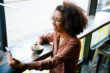 Kapalı kafede cep telefonu ve kulaklık kullanan afro saçlı genç siyahi bir kadın.